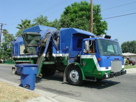 A large rubbish removal truck with a predominantly blue and green exterior is parked on a suburban street, surrounded by greenery including a large tree and some palm trees in the background. The truck has a front cab with a metallic silver grille and a curved windshield, and is fitted with a hydraulic lifting mechanism at the rear, which is actively engaged with a dark green waste bin positioned at the back left side of the truck. The waste bin has a textured surface with a lid that is slightly open, and shows signs of use. The truck’s body features reflective yellow and orange markings, and the scene is illuminated by natural daylight, indicating clear weather conditions. The area around the truck includes a paved road, a grassy verge, and a residential setting with a visible row of houses and electrical poles in the background. This image, associated with Garden Clearance Elephant and Castle’s rubbish services, highlights the process of waste collection in a quiet, well-maintained neighborhood in south London or nearby postcode areas.