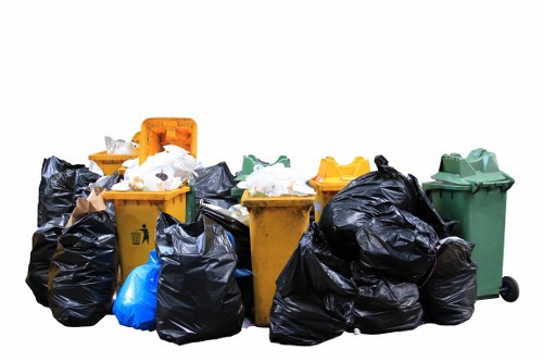 A collection of various refuse bins and garbage bags positioned outdoors on a paved surface, with some bins slightly in front of others. The bins include a yellow one, a green one, and an orange one, all with closed lids, and are made of plastic with a smooth finish and visible wheels on the green bin. The garbage bags are predominantly black, with a few blue ones, and are filled with mixed waste, some of which appear to be loosely tied or spilling slightly at the top. The scene appears to be set outside a property in London, as suggested by the context of garden clearance services in Elephant and Castle. The background is plain and white, emphasizing the refuse collection, which relates to rubbish removal services. The lighting is natural, with even illumination across the objects. The image is relevant for a rubbish removal website, highlighting waste disposal and clearance services in a residential or commercial setting in the London area, specifically near the postcode zone of Elephant and Castle.