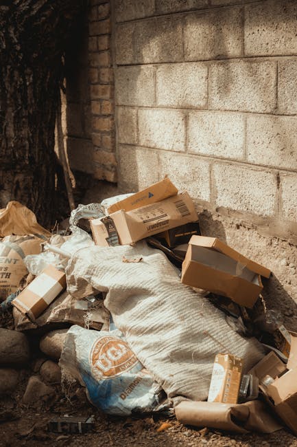 A large accumulation of mixed household waste and rubbish bags is piled on a paved street near a parking area, with some cardboard boxes and discarded packaging scattered across the ground. The rubbis