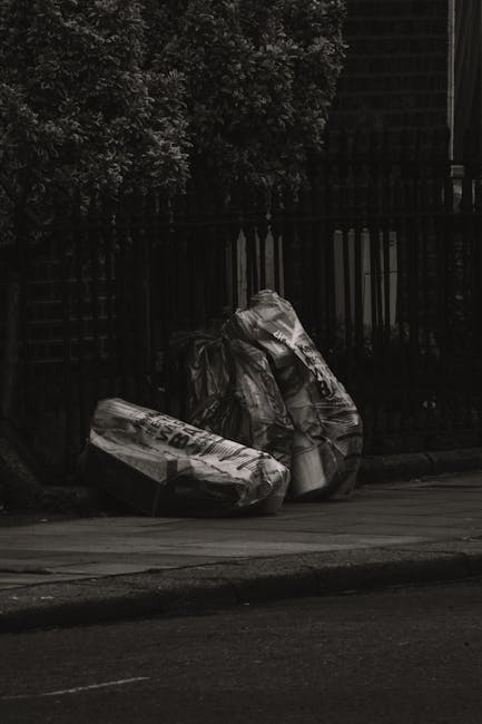 Two large black plastic rubbish bags, tightly packed and leaning against a black metal fence on a street pavement, with each bag appearing to contain mixed waste materials such as paper, packaging, an