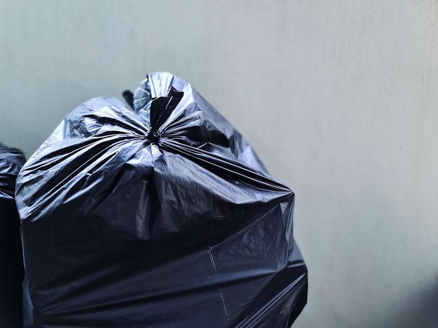 Two large black plastic rubbish bags, tightly packed and leaning against a black metal fence on a street pavement, with each bag appearing to contain mixed waste materials such as paper, packaging, an